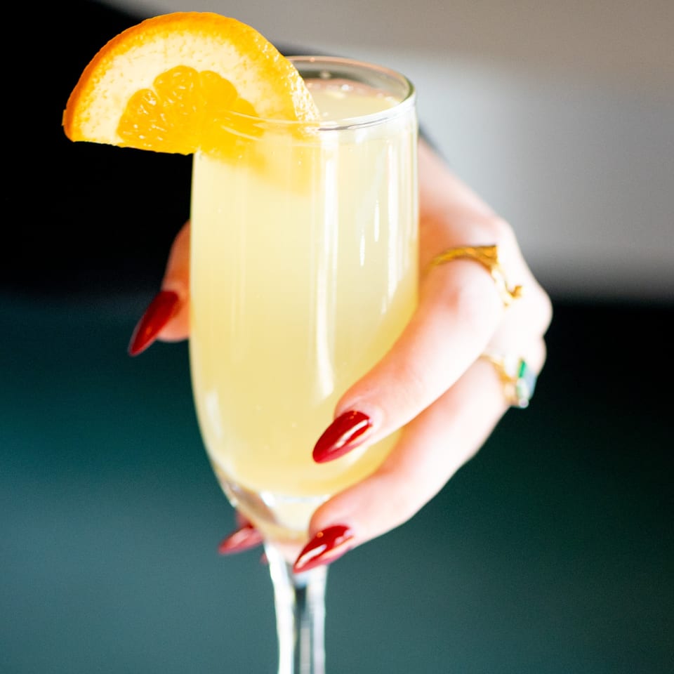 A hand with red manicured nails holding a mimosa garnished with an orange slice.