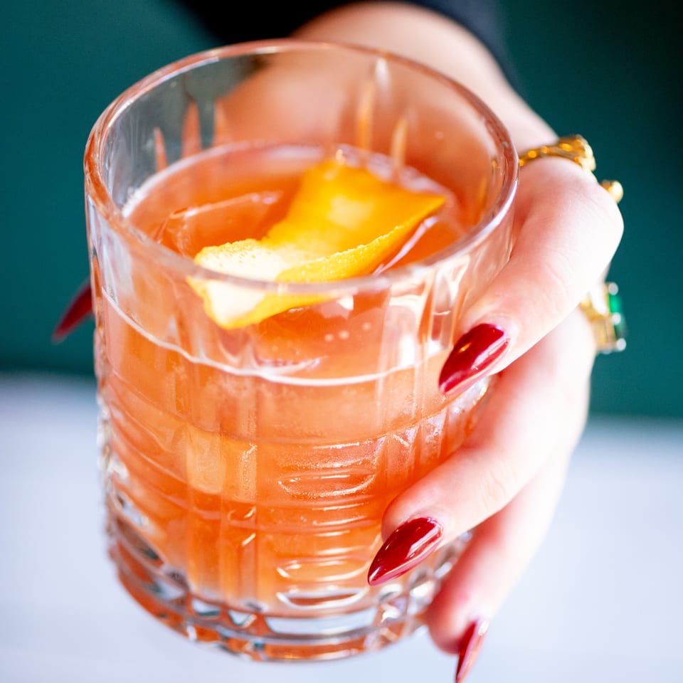 A hand with red nails holds a glass of orange cocktail garnished with an orange peel.