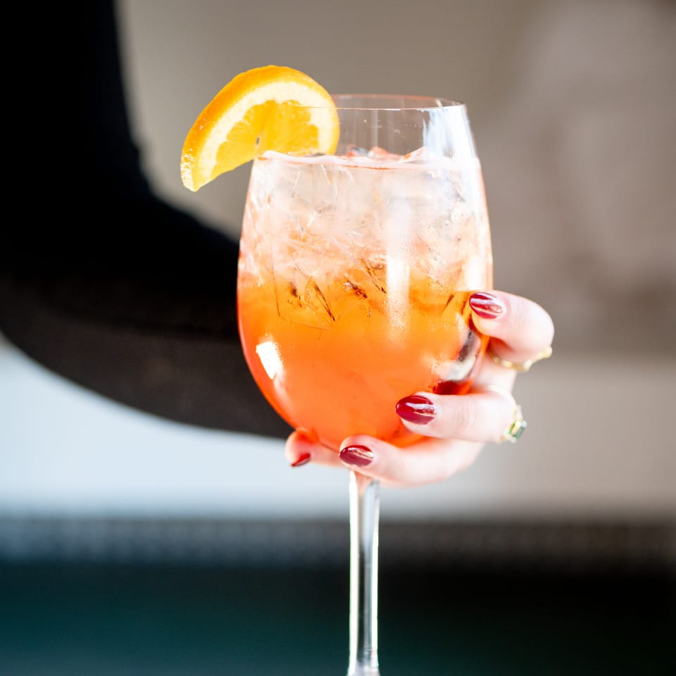 A hand with red manicured nails holding a vibrant orange Aperol Spritz cocktail with an orange slice.