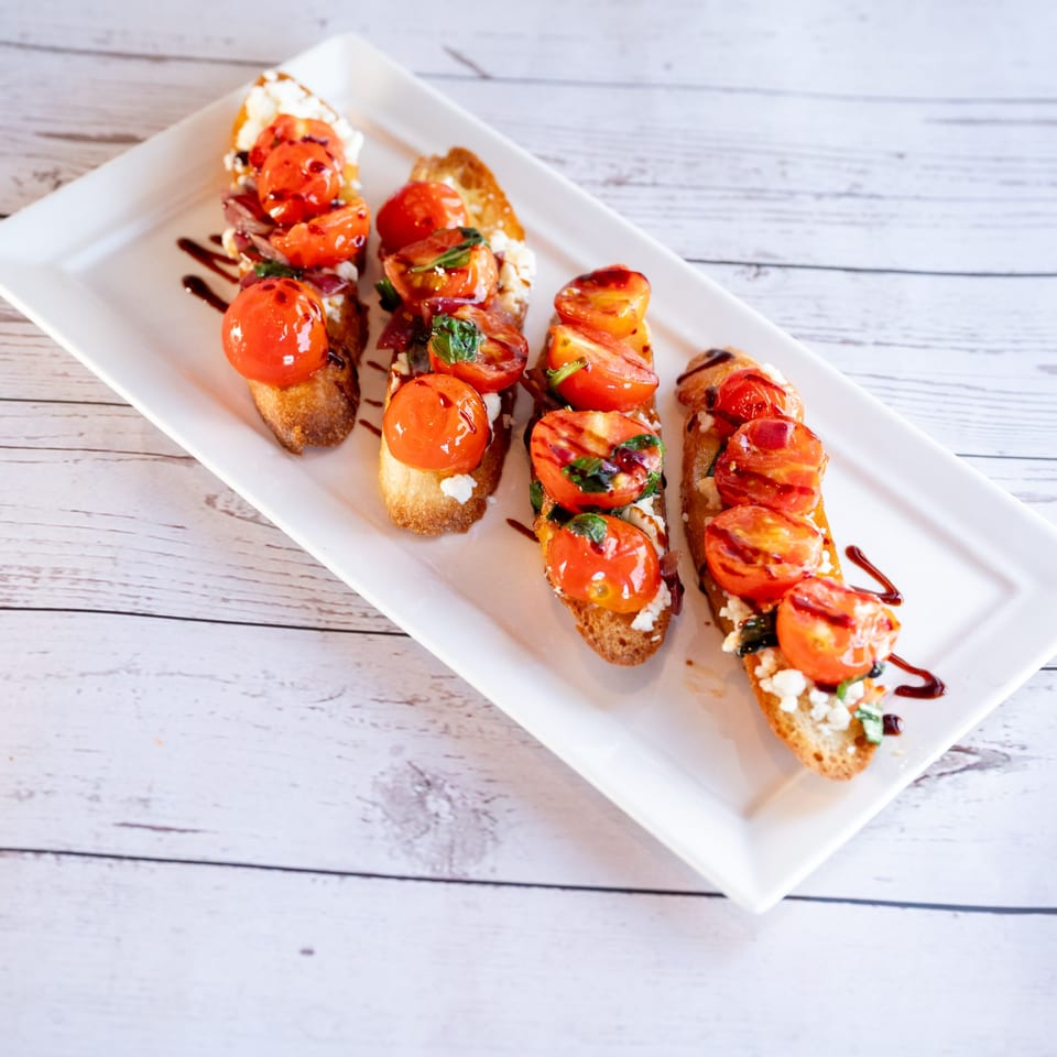 Four bruschetta appetizers with roasted tomatoes, cheese, and balsamic glaze on a white rectangular plate.