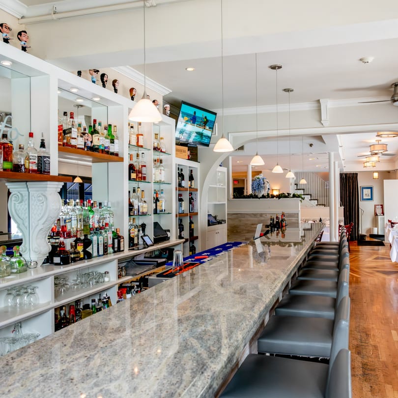 Long, well-stocked bar with a granite top and gray stools, leading to a dining area.