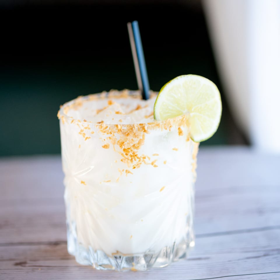 A white cocktail with a toasted coconut rim, lime wedge, and blue straw on a wooden table.